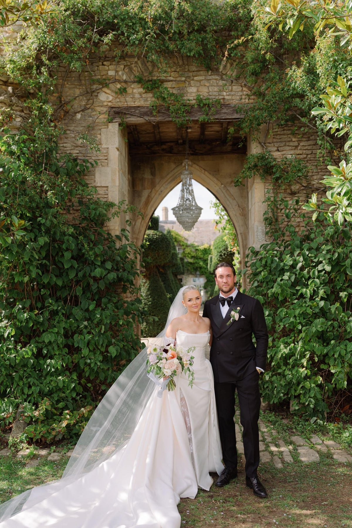 Bride and groom beneath a stone arch, with the groom wearing black tie tailoring